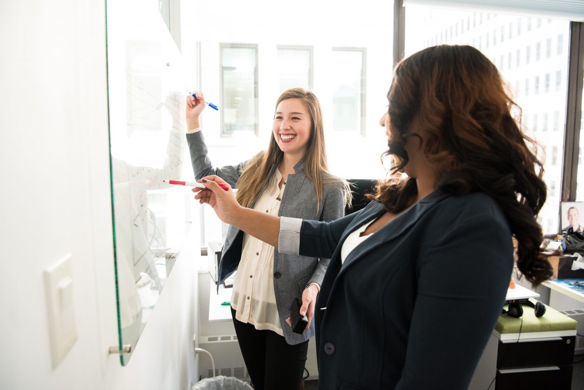 Two female employees smiling and writing on a white board in the office